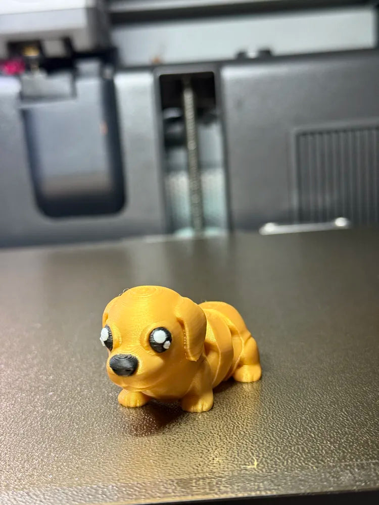 Cute yellow 3D printed Flexi Mini Labrador by Wiggle Nest, featuring large black-and-white eyes, floppy ears, black nose and an articulated body, displayed on a warm studio-style wooden background.