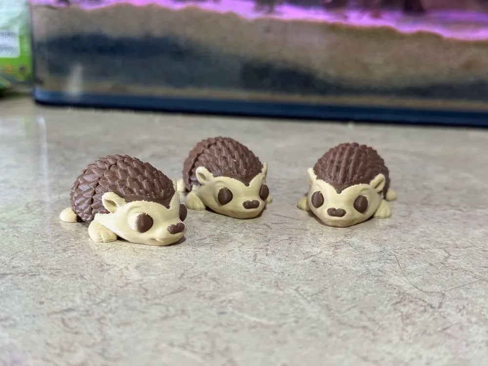 Three cute Flexi Mini Hedgehog 3D-printed toys in cream and brown, displayed on a warm wooden tabletop with plants and a cozy natural background. Wigglenest collectible articulated mini toys designed for playful display and gifting