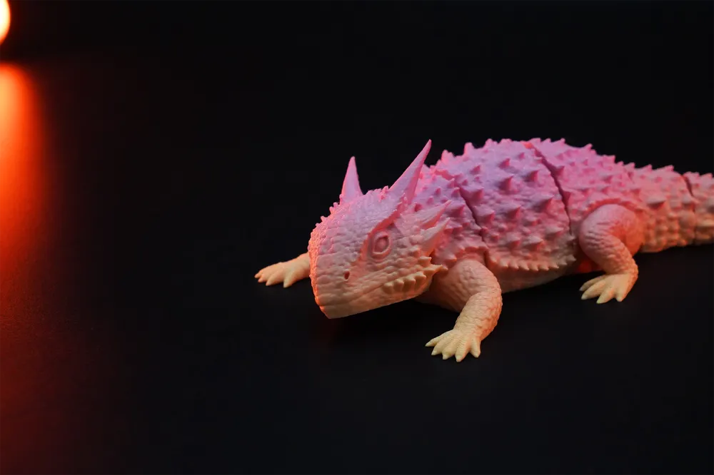 Three pastel yellow and light blue 3D-printed Flexi Horned Lizard collectibles displayed in a warm nature-inspired setting with rocks, greenery and wooden accents. The articulated lizards feature detailed textured scales, horns and flexible segmented bodies, presented as unique PLA collectibles by Wigglenest.
