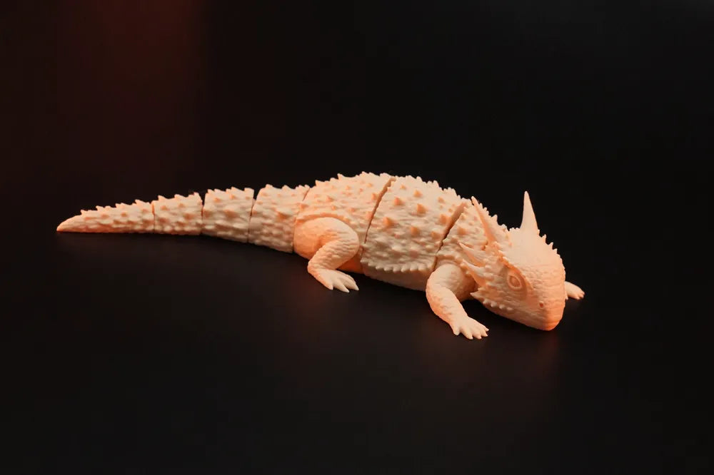 Three pastel yellow and light blue 3D-printed Flexi Horned Lizard collectibles displayed in a warm nature-inspired setting with rocks, greenery and wooden accents. The articulated lizards feature detailed textured scales, horns and flexible segmented bodies, presented as unique PLA collectibles by Wigglenest.