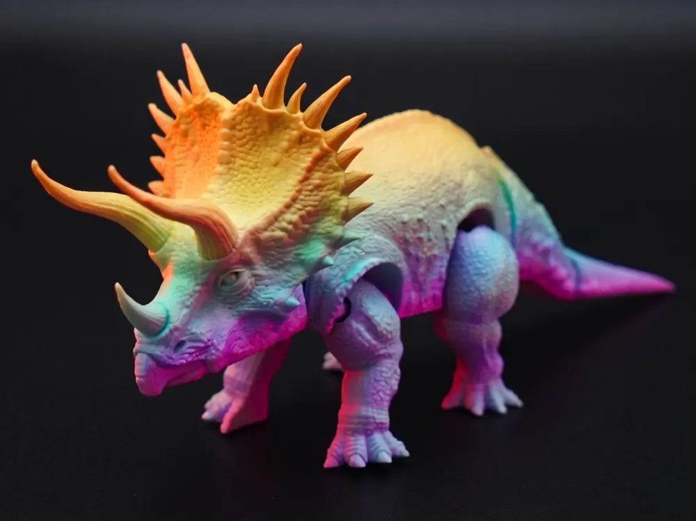Colorful 3D-printed articulated Triceratops collectible featuring detailed horns and textured skin, displayed on a rocky prehistoric-style setting with dramatic warm lighting and a softly blurred natural background