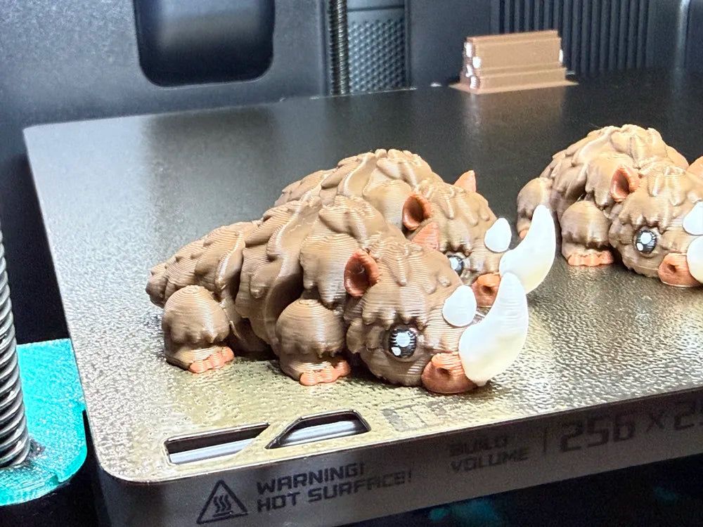 Flexi Mini Woolly Rhino 3D-printed collectibles with cute brown textured bodies, large white horns and articulated sections, displayed in a snowy mountain scene at sunset.