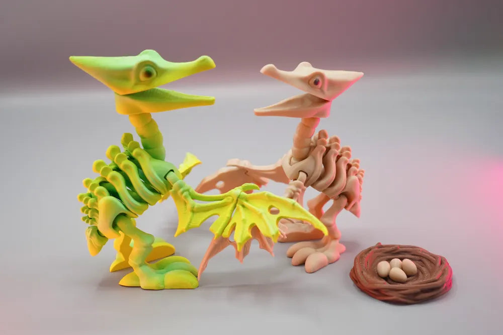 Two 3D printed articulated pterosaur skeleton toys in green and natural bone colours posed together beside a miniature nest with eggs.